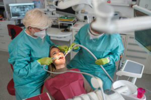 While Caucasian female dentist working with young patient female dental assistant helping around. Both wearing a protective gowns, face masks and gloves during covid-19 pandemic.