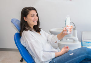 A young woman who might have had a dental plant is looking at a mirror with a smile In a Dentist's office.