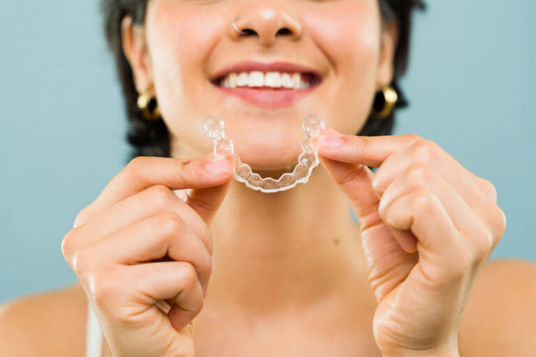 A close-up image of a smiling young woman holding invisible braces for teeth alignment.