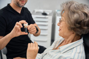 A senior woman patient examines a model of a dental implant in the hands of a dental prosthetist.