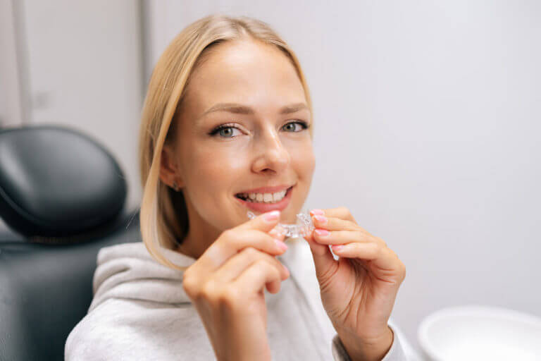 A close-up portrait of a smiling blonde female patient holding an invisible braces aligner while sitting in a dentistry clinic.